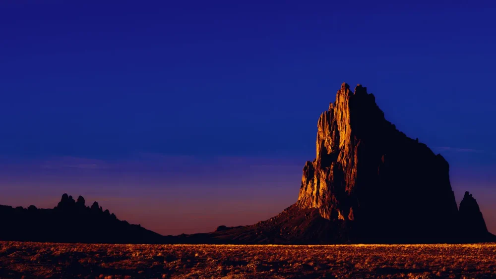 The volcanic neck of Shiprock (Tsé Bitʼaʼí) rises 1,583 feet above the surrounding desert plain, lit by the last warm light of the day against a deep indigo sky. Navajo Nation, northwestern New Mexico. Fine art landscape photograph.