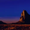 The volcanic neck of Shiprock (Tsé Bitʼaʼí) rises 1,583 feet above the surrounding desert plain, lit by the last warm light of the day against a deep indigo sky. Navajo Nation, northwestern New Mexico. Fine art landscape photograph.