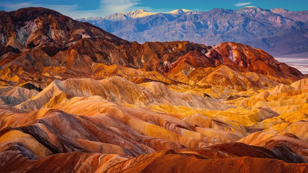 Golden and rust-colored eroded badlands of Zabriskie Point rise against distant Panamint Mountains at first light, Death Valley National Park, California. Fine art landscape photograph.