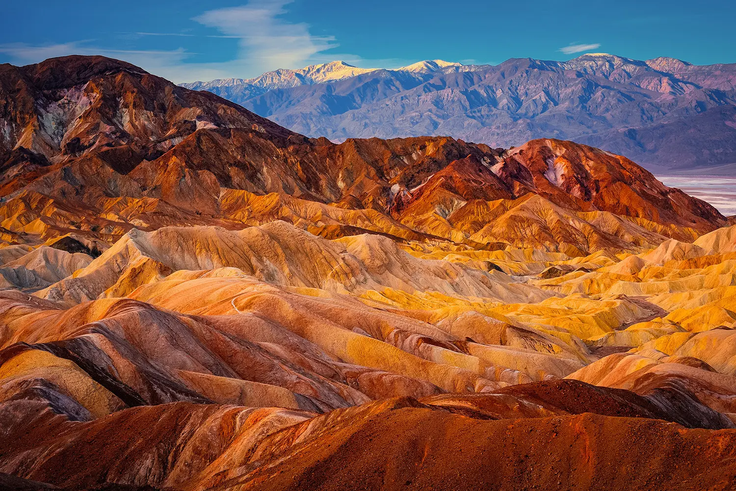 Golden and rust-colored eroded badlands of Zabriskie Point rise against distant Panamint Mountains at first light, Death Valley National Park, California. Fine art landscape photograph.