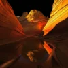 The swirling cross-bedded sandstone of the Wave at Coyote Buttes North ignites in warm afternoon light with a rare ephemeral water reflection doubling the composition below. Vermilion Cliffs National Monument, Arizona. Fine art landscape photograph. Fujifilm GFX medium format.