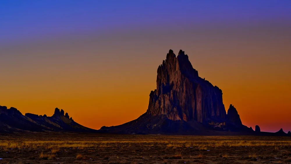 The volcanic neck of Shiprock (Tsé Bitʼaʼí) stands in complete silhouette against a burning twilight gradient sky, deep cobalt transitioning through amber to burning orange at the horizon. Navajo Nation, northwestern New Mexico. Fine art landscape photograph. Nikon medium format