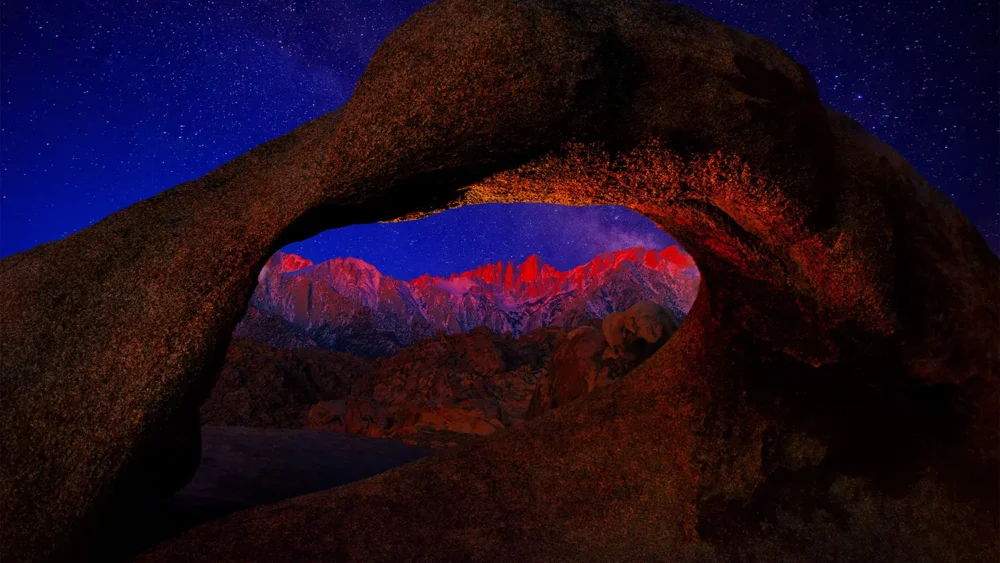 Mobius Arch in the Alabama Hills silhouetted against a starry night sky, framing the distant Sierra Nevada peaks illuminated in pink alpenglow at first light. Alabama Hills, Inyo County, California. Fine art landscape photograph with astrophotography composition. Nikon medium format