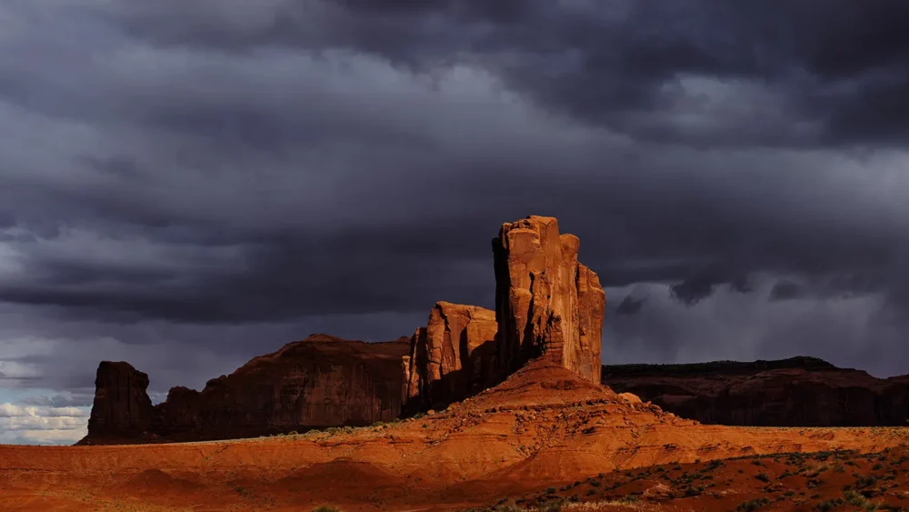 Red sandstone buttes of Monument Valley illuminated by a break in dark storm clouds while the surrounding desert remains in shadow, Navajo Tribal Park, Arizona-Utah border. Fine art landscape photograph. Fujifilm GFX medium format.