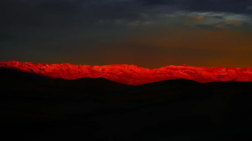 The Panamint Range ignites in deep crimson alpenglow at sunset, viewed across the dark floor of Death Valley, California. Panoramic fine art landscape photograph. Hasselblad X1D II medium format.