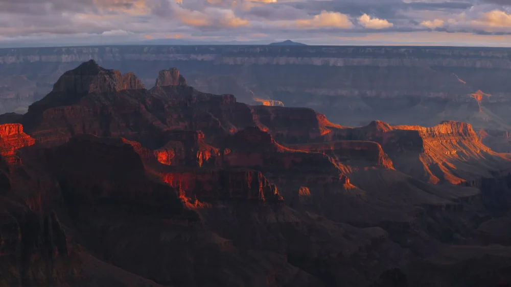 The Grand Canyon from the North Rim ignites into burning orange and red at the specific atmospheric moment when direct sunset light strikes the canyon walls, deep purple shadow filling the canyon depths and dramatic cloud shadows above. Arizona. Panoramic fine art landscape photograph. Fujifilm GFX medium format.