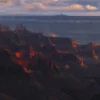 The Grand Canyon from the North Rim ignites into burning orange and red at the specific atmospheric moment when direct sunset light strikes the canyon walls, deep purple shadow filling the canyon depths and dramatic cloud shadows above. Arizona. Panoramic fine art landscape photograph. Fujifilm GFX medium format.