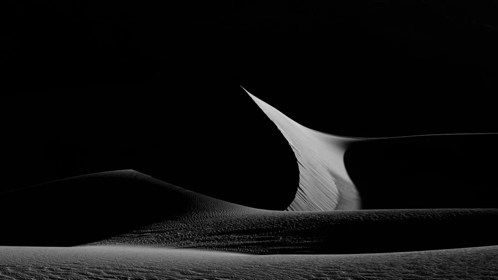 A single curved dune ridge lit by sidelight against pure black shadow at Eureka Dunes, Death Valley National Park, California. Black and white fine art landscape photograph. Fujifilm GFX