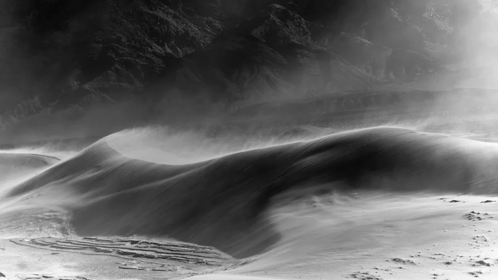 Wind-shaped dunes with visible sand movement and distant ridgeline at Eureka Dunes, Death Valley National Park. Black and white atmospheric fine art landscape photograph. Fujifilm GFX medium format