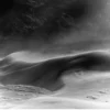 Wind-shaped dunes with visible sand movement and distant ridgeline at Eureka Dunes, Death Valley National Park. Black and white atmospheric fine art landscape photograph. Fujifilm GFX medium format