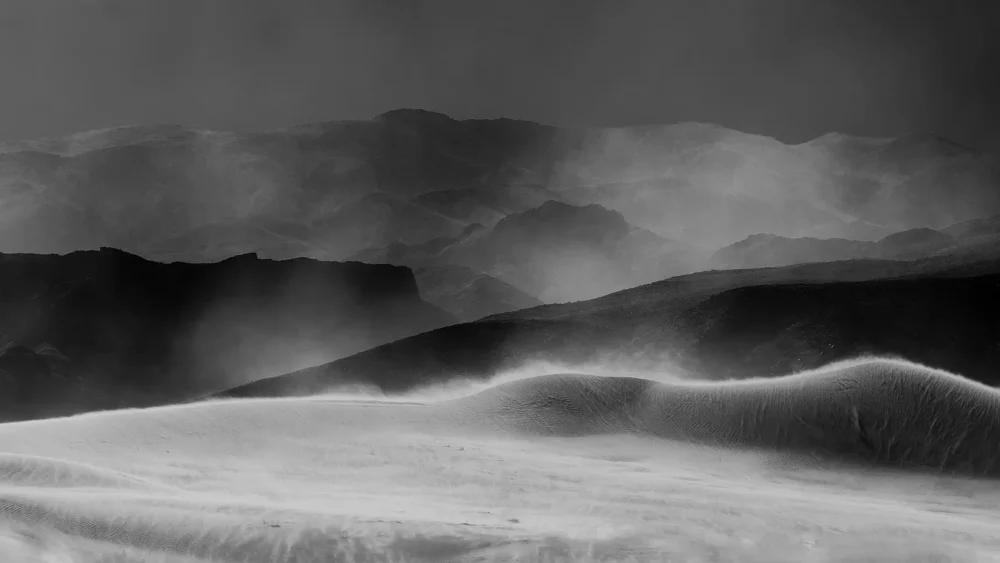 Layered dune forms with visible wind-drift patterns and distant ridgeline at Eureka Dunes, Death Valley National Park. Black and white atmospheric fine art landscape photograph. Fujifilm GFX medium format