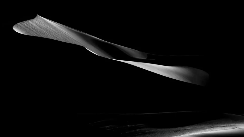 A single sculptural dune form catches sidelight and appears suspended against pure black shadow at Eureka Dunes, Death Valley National Park. Black and white minimalist fine art photograph. Fujifilm GFX medium format.