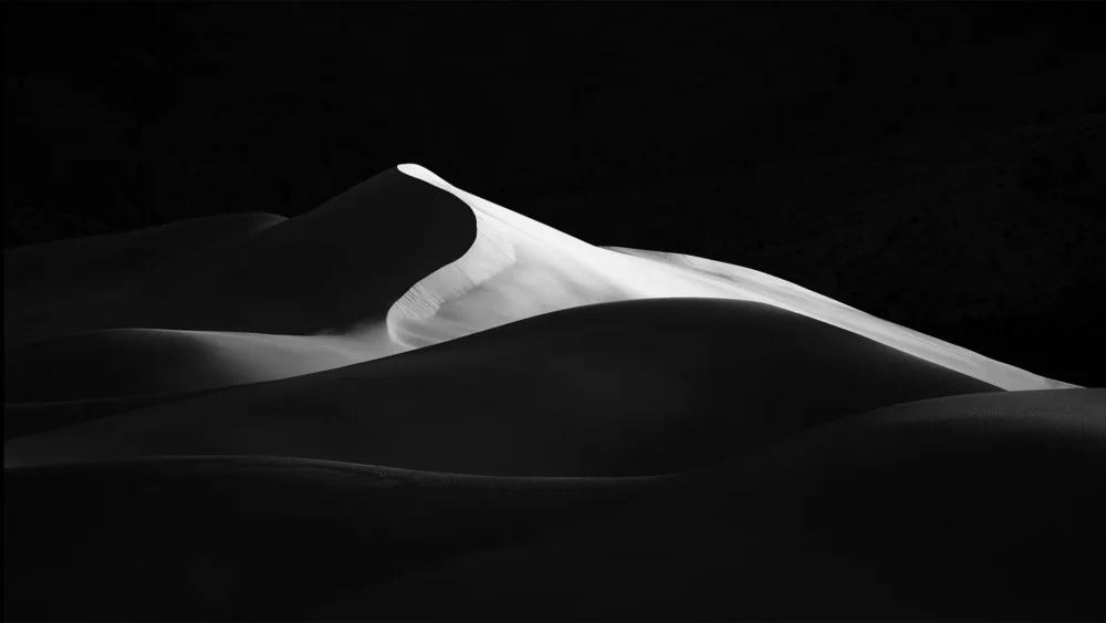 A single luminous white dune ridge rises against pure black shadow at Eureka Dunes, Death Valley National Park. Panoramic black and white fine art photograph. Fujifilm GFX medium format.