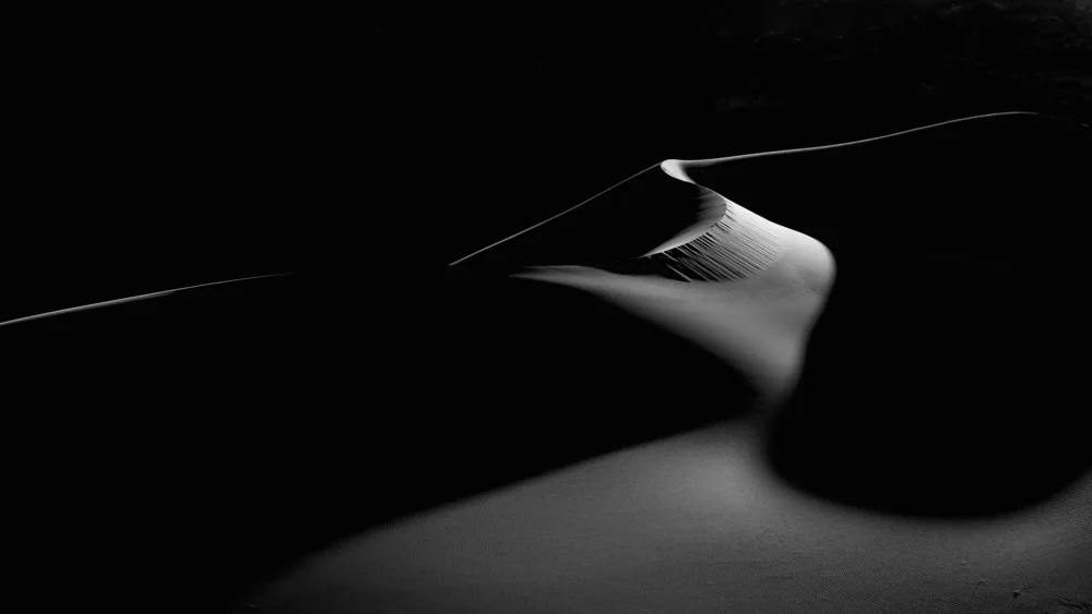 A single white dune ridge sweeps across pure black shadow like a cresting wave at Eureka Dunes, Death Valley National Park. Black and white panoramic fine art photograph. Fujifilm GFX medium format
